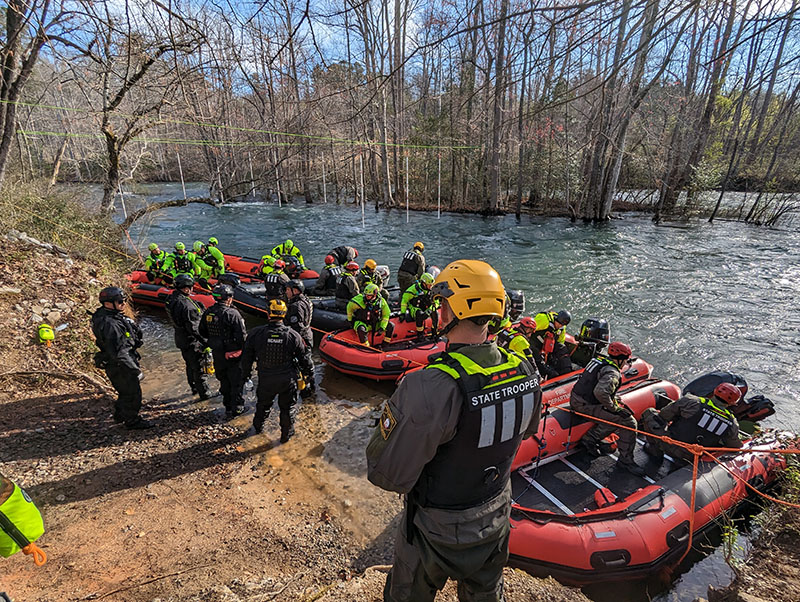 Students training in a swiftwater rescue class board boats in preparation for learning best practices in saving drowning victims in swiftwater situations, particularly during flooding or other treacherous situations on swollen rivers, lakes and streams.